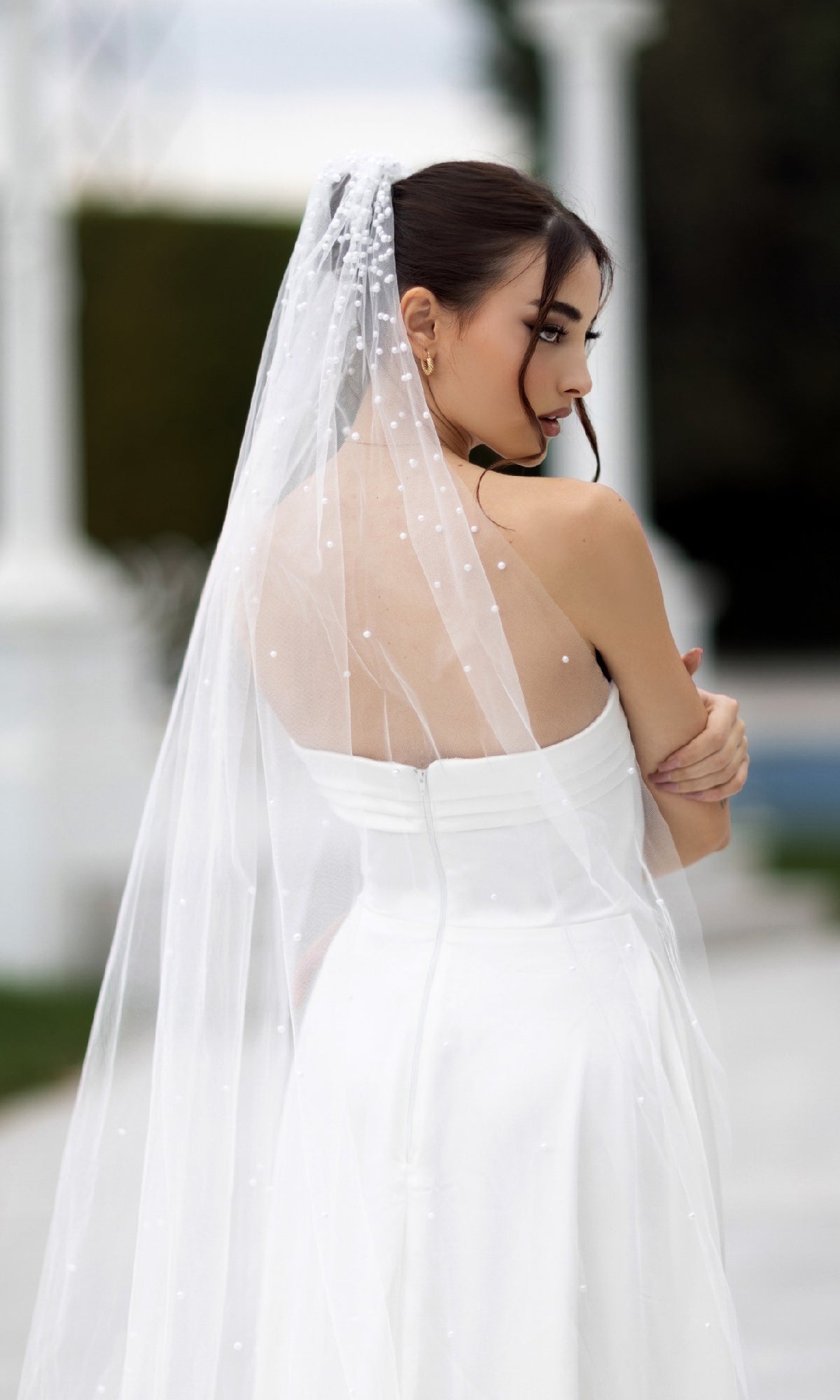 A woman in the Off-White Strapless Bridal Gown: Tina Holly TW105 and a sheer veil stands outdoors, gazing gracefully over her shoulder.